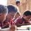 A group of Orang Asli Bateq children engaged in writing and drawing activities in an outdoor classroom.