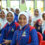 A group of cheerful young girls in school uniforms, smiling and making peace signs during an educational program in a school hall.