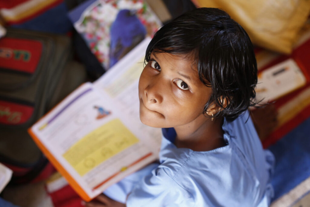 A young girl in India holding a book looks up to the camera.